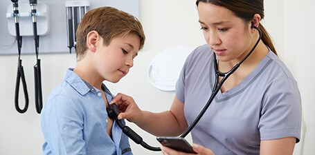 Clinicien auscultant la poitrine d’un garçon tout en regardant une application sur téléphone
